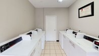 This single-floor shared laundry facility is designed for practicality and efficiency, offering a clean and organized environment for laundry needs. The room is finished with neutral gray walls and light-toned square floor tiles that contribute to a bright, tidy aesthetic throughout.

Multiple top-loading washing machines and dryers are arranged along the walls on both sides of the room, providing ample capacity for simultaneous use. A centrally positioned folding table with a white surface and metal frame offers a convenient workspace for sorting and folding laundry. Additional built-in utility features, including recessed wall compartments with plumbing hookups and standard electrical outlets, are distributed throughout the space for added functionality.

A coin-operated payment machine is stationed near the entrance, streamlining the self-service experience. Safety is addressed with a prominently mounted fire extinguisher. A framed notice or certificate is displayed on the wall, adding a finished touch to the space.

Natural light enters through a side window fitted with horizontal blinds, complementing the overhead ceiling light fixture. A white paneled entry door with a glass insert provides access to the exterior, allowing for ventilation and a visual connection to the outside. The overall ambiance is clean, utilitarian, and well-maintained, making it a practical and accessible communal laundry space.