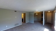 This single-floor space offers a straightforward and functional layout designed for comfortable everyday living. The main living area features wall-to-wall carpeting in a neutral tone, providing a soft and cohesive foundation throughout the space. The walls are painted in a calm gray hue, creating a clean, understated backdrop that suits a variety of furnishing styles.

A textured popcorn ceiling spans the living area, and floor-level air vents are positioned along the walls, indicating a central HVAC system. A thermostat is also visible on the wall, adding to the practical utility of the space.

Adjacent to the living area, a dining space is defined by a hanging chandelier-style light fixture, offering a designated area for meals. This area flows naturally toward the kitchen, which is partially visible and appears to include cabinetry along the walls.

A doorway from the main living space leads into a secondary room, warmly lit and providing access to what appears to be a bathroom or hallway. This interconnected layout supports easy movement between areas.

Overall, the space presents a practical and neutral interior with an open feel, plentiful floor space, and a connected layout that links the living, dining, and kitchen areas efficiently.