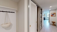This single-floor unit offers a thoughtfully designed layout that balances functionality with contemporary style. The living area is bright and welcoming, featuring light hardwood flooring, clean white walls, and large windows that frame urban cityscape views, filling the space with natural light. A colorful area rug and eclectic furnishings add personality to the open-plan design, which flows seamlessly between the living and sleeping areas.

The kitchen is efficiently arranged with light wood cabinetry, stone-finish countertops, and a striking iridescent vertical tile backsplash. Stainless steel appliances, open shelving, and a window overlooking the city skyline complete this well-equipped cooking space.

The bedroom is spacious and serene, finished with the same light hardwood flooring. It features a sliding closet and direct access to an en-suite bathroom. The bathroom is finished with black-and-white patterned floor tiles and a frameless glass sliding shower enclosure, delivering a clean, modern aesthetic.

A dedicated entryway features a built-in bench with drawers and a row of wall hooks, providing practical and organized storage. Additional hallway storage is provided by louvered sliding closet doors revealing built-in shelving, maximizing the unit's overall storage capacity.
