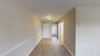 This single-floor unit offers a practical and well-organized layout with a neutral color palette throughout. The main living area features light wood-look flooring, warm beige walls, white baseboards, and a textured ceiling, creating a clean, understated aesthetic. A hallway connects the primary spaces, with doors leading to additional rooms and natural light filtering in through a window at the far end.

The kitchen is efficiently designed, featuring white cabinetry with sleek bar-pull hardware, light granite-style countertops, a stainless steel sink, and black appliances including a refrigerator, electric range, and range hood. The layout maximizes storage and counter workspace effectively.

The bedrooms are carpeted and simply finished, each offering a calm, neutral environment. One bedroom includes a closet with a six-panel white door, while another room is outfitted with wood-look flooring and similarly clean finishes. A standout feature is the room with a ceiling fan and a large sliding glass door with vertical blinds, providing access to an outdoor area and allowing generous natural light.

The bathroom is neatly appointed with a single-sink vanity featuring a granite-style top, a full tub-shower combination with white tile surround, and a textured accent wall, rounding out this straightforward and functional space.