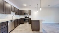 This single-floor unit offers a well-designed layout with a clean, contemporary aesthetic throughout. The kitchen serves as a central highlight, showcasing rich dark-toned shaker-style cabinetry paired with granite countertops and a white subway tile backsplash. A full suite of stainless steel appliances includes a refrigerator, range, microwave, and dishwasher. Pendant lighting fixtures hang above a kitchen island that provides additional counter and prep space, while light wood-look flooring runs throughout the main living areas, creating a bright and cohesive look.

The open floor plan connects the kitchen to the living area, which benefits from a sliding glass door that introduces natural light and outdoor access. A ceiling fan adds practical comfort to the space. The bedroom is finished with plush carpet and a ceiling fan, offering a quiet, comfortable retreat. It connects to a spacious walk-in closet equipped with built-in shelving and rod storage along multiple walls, providing generous organizational capacity.

The bathroom features a dark-toned vanity with a granite countertop, a tiled shower-tub combination, and the same light wood-look flooring found throughout the unit. The overall palette of white walls, dark cabinetry, and light flooring creates a modern, move-in-ready environment.