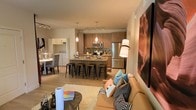 This thoughtfully designed single-floor residence showcases a contemporary open-concept layout that seamlessly connects the living, dining, and kitchen areas. The living space is anchored by a comfortable tan sofa adorned with colorful accent pillows and is elevated by striking large-format canvas artwork depicting vivid canyon landscapes, adding a bold visual focal point to the room. A floor lamp provides warm ambient lighting, complementing the flush ceiling light fixture overhead. The kitchen is a standout feature, boasting sleek wood-toned cabinetry, stainless steel appliances including a refrigerator, microwave, and range, as well as a stone-topped island with bar seating for four — ideal for casual dining or entertaining. Pendant lighting above the island adds a modern, polished touch, while track lighting illuminates the full length of the kitchen. Light wood-toned flooring runs throughout, contributing to the space's bright and cohesive aesthetic. A dining area with upholstered chairs sits adjacent to the kitchen, and a washer and dryer are conveniently situated nearby. A glass-paneled entry door allows natural light to filter into the living area, enhancing the overall openness and airy feel of the space.