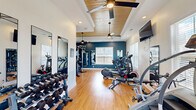 Located at 874 Needmore Rd in Clarksville, Tennessee, this fitness center offers a thoughtfully designed exercise space on a single floor. The room is defined by its standout architectural detail: a tray ceiling accented with warm wood paneling and fitted with dual ceiling fans, lending both style and comfort to the workout environment. Recessed lighting complements the abundant natural light that streams in through multiple windows dressed with white blinds, creating a bright and energizing atmosphere.

The flooring throughout is polished hardwood, providing a clean and durable surface suited to a variety of workouts. The walls are painted in a bold deep teal on one side and crisp white on the other, giving the space a modern, motivating aesthetic. Full-length mirrors are mounted along the walls, enhancing the sense of openness and allowing users to monitor their form.

The exercise room is comprehensively equipped for both cardio and strength training. Cardio equipment includes treadmills, elliptical machines, a stationary bike, and a stair climber. The strength training area features a multi-station cable machine, a weight bench, barbells, a punching bag, kettlebells, and a dumbbell rack. A wall-mounted television adds an entertainment option during workouts. A glass entry door provides direct exterior access, adding convenience for users.