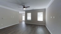 Located at 3112 Maybank Hwy on scenic Johns Island, South Carolina, this single-floor home offers a welcoming and functional layout designed for comfortable everyday living. The interior is finished with rich, dark hardwood-style flooring that runs throughout, providing a warm and cohesive aesthetic that complements the neutral gray-toned walls. Crown molding adds a refined architectural detail, lending the space a polished, well-crafted feel. The main living area is generously proportioned, with multiple windows dressed in white horizontal blinds that allow natural light to filter in while maintaining privacy. A ceiling fan with a built-in light fixture keeps the space comfortable year-round. The open sightlines connect the living area toward what appears to be an entry or transitional space, enhancing the sense of flow and openness throughout the home. Clean white baseboards provide crisp contrast against the darker flooring, and the overall color palette creates a calm, move-in-ready environment. The single-story layout ensures ease of accessibility throughout, making this Johns Island property a straightforward and appealing option for those seeking a well-maintained, simply designed home in the greater Charleston area.