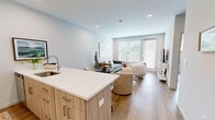 This thoughtfully laid-out one-bedroom, one-bathroom residence offers a bright and modern living experience across a single floor. The open-concept kitchen and living area serves as the heart of the home, where light wood cabinetry, white quartz countertops, and stainless steel appliances create a clean, contemporary kitchen. A kitchen island with an undermount sink adds both functionality and style, flowing seamlessly into the living area. The living room is bathed in natural light from large windows overlooking greenery, with wood-look flooring that runs throughout the space, lending warmth and continuity. Recessed lighting and soft, neutral wall tones contribute to the airy, welcoming ambiance. The bedroom is a calm retreat, featuring a window with wooded views, wood-look flooring, and a closet for ample storage. A hallway connects the main living areas to the private spaces. The bathroom is finished in crisp white tones, featuring a tub-shower combination with built-in shelving, wood-look flooring, and clean, minimal fixtures. Overall, this residence presents a cohesive modern aesthetic with functional design, making efficient use of its well-organized layout.