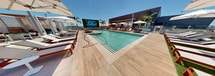 This upscale outdoor recreational space showcases a stunning resort-style pool area designed for relaxation and entertainment. The centerpiece is a large, L-shaped swimming pool with crystal-clear turquoise water, complemented by a separate lap pool and a smaller wading pool with built-in steps. The expansive deck area is finished with high-quality materials, featuring a mix of tile and wood textures that add warmth and sophistication.

Comfortable sun loungers line the pools, offering ample seating for sunbathing and relaxation. White umbrellas provide shade options, while private cabanas with louvered sides offer a more secluded experience. Palm trees dot the landscape, enhancing the tropical resort feel.

The modern architectural design of the adjacent building features clean lines and large glass walls, allowing for a seamless indoor-outdoor flow. A lush living wall and strategically placed planters add greenery to the space. The area is well-lit with contemporary light fixtures, ensuring the space can be enjoyed day and night.

This meticulously designed outdoor area creates an oasis-like atmosphere, perfect for those seeking a luxury resort experience without leaving the premises. The thoughtful layout and high-end amenities cater to both active lifestyles and leisurely relaxation, making it an ideal space for social gatherings or personal rejuvenation.