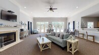 Located at 1878 Gingercake Cir in Rock Hill, SC, this single-story community clubhouse is designed to provide a welcoming and functional gathering space for residents. The interior is finished with soft gray walls, crown molding, and plush carpeting throughout the main areas, creating a clean and cohesive aesthetic.

The spacious main lounge serves as the heart of the facility, featuring a classic white-mantled fireplace with a wall-mounted flat-screen television above, ceiling fans, and recessed lighting. Comfortable seating arrangements and rustic wood accent tables fill the room, while large French doors along the rear wall flood the space with natural light and provide direct views and access to an outdoor pool area.

The entry reception zone is defined by elegant white columns, tile flooring, and an open layout that connects seamlessly to the lounge. A service counter and informational displays are conveniently positioned near the entrance for resident use.

A kitchenette area with light wood cabinetry, a breakfast bar with seating, and standard appliances offers a practical amenity for gatherings and events. Adjacent seating with tufted armchairs provides a relaxed nook for conversation.

Throughout the space, a navy blue and coastal-inspired décor theme ties together the furnishings and artwork, giving the clubhouse an inviting, resort-like ambiance suited for both everyday use and community events.