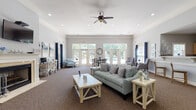 Located at 1878 Gingercake Cir in Rock Hill, SC, this single-story community clubhouse is designed to provide a welcoming and functional gathering space for residents. The interior is finished with soft gray walls, crown molding, and plush carpeting throughout the main areas, creating a clean and cohesive aesthetic.

The spacious main lounge serves as the heart of the facility, featuring a classic white-mantled fireplace with a wall-mounted flat-screen television above, ceiling fans, and recessed lighting. Comfortable seating arrangements and rustic wood accent tables fill the room, while large French doors along the rear wall flood the space with natural light and provide direct views and access to an outdoor pool area.

The entry reception zone is defined by elegant white columns, tile flooring, and an open layout that connects seamlessly to the lounge. A service counter and informational displays are conveniently positioned near the entrance for resident use.

A kitchenette area with light wood cabinetry, a breakfast bar with seating, and standard appliances offers a practical amenity for gatherings and events. Adjacent seating with tufted armchairs provides a relaxed nook for conversation.

Throughout the space, a navy blue and coastal-inspired décor theme ties together the furnishings and artwork, giving the clubhouse an inviting, resort-like ambiance suited for both everyday use and community events.