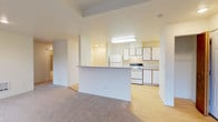 This single-floor unit at 13107 East Mission Avenue in Spokane, Washington is designed with a practical, open layout that connects the living and kitchen areas seamlessly. The kitchen features light wood-tone flooring, white cabinetry with oak-trimmed accents, a white electric range, a full-size refrigerator, and ample counter space. A built-in desk nook provides a functional workspace just off the kitchen. A half-wall peninsula separates the kitchen from the carpeted living area, which benefits from natural light and direct access to a private balcony through a sliding glass door. The balcony overlooks a tree-lined exterior, providing a pleasant outdoor view. A wall-mounted air conditioning unit and a baseboard heater serve the living space for year-round comfort. The bedrooms are carpeted and well-lit by multiple windows with views of surrounding trees. One bedroom includes a sliding wood-door closet with built-in shelving for organized storage. The bathroom is finished with tile flooring, a wide vanity with a large mirror, a soaking tub, and a frosted glass block window for natural light and privacy. A coat closet is conveniently located near the entry.