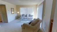 This single-floor space offers a warm and inviting atmosphere, highlighted by a well-appointed living room that serves as the heart of the home. The living room features soft, wall-to-wall carpeting in a neutral tone, creating a comfortable and cohesive foundation throughout the space. Large windows dressed with vertical blinds allow generous amounts of natural light to filter in, brightening the room and lending it an airy, open feel. The walls are finished in a soft, warm beige, complementing the overall light and neutral color palette. A textured ceiling adds subtle visual interest overhead. The living area comfortably accommodates generous seating, including a plush sofa adorned with decorative accent pillows and a matching armchair, all arranged around a sleek glass-top coffee table that adds a contemporary touch. A glass side table with a standing lamp provides additional ambiance and functionality. The space is thoughtfully designed for relaxed, everyday living, with a clean layout that allows for easy movement and a sense of openness. Overall, the space presents a calm, neutral aesthetic that is both functional and welcoming.
