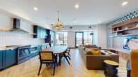 This thoughtfully designed single-floor space seamlessly integrates a kitchen, dining area, and living room into one expansive open-plan layout. The kitchen is a standout feature, showcasing bold teal shaker-style cabinetry with brushed gold circular hardware across both upper and lower units. White square-tile backsplash, light quartz countertops, stainless steel appliances including a range, dishwasher, and built-in microwave, and a sleek range hood complete the well-appointed cooking space.

The dining area sits centrally, anchored by a solid wood oval table surrounded by cane-back chairs beneath a sculptural brass multi-pendant chandelier. This zone flows naturally into the living area, where a generous sectional sofa in warm brown tones is paired with mustard accent cushions. A dramatic arc floor lamp with a matte navy shade adds character, while a built-in walnut shelving unit with geometric-patterned wallpaper backing frames the television wall.

Throughout the space, light herringbone-patterned hardwood flooring ties the zones together cohesively. Floor-to-ceiling windows and glass sliding doors flood the interior with natural light and provide direct access to an outdoor terrace with open views, enhancing the overall sense of openness and connectivity to the outdoors.