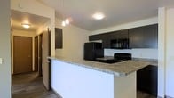 This single-floor space showcases a thoughtfully designed open-concept layout that seamlessly connects the kitchen to the living areas. The kitchen serves as the focal point, featuring bold dark cabinetry that provides a striking contrast against the light-toned walls. A spacious peninsula with granite-style countertops offers both additional prep space and a natural gathering point, making the kitchen as functional as it is visually appealing. Black appliances — including a refrigerator, range, and built-in microwave — lend a cohesive, contemporary aesthetic throughout the space. Overhead, a set of modern pendant lights hangs above the peninsula, providing targeted task lighting while adding a decorative touch. A flush-mount ceiling fixture contributes to the overall brightness of the room. The flooring features wood-look planks that add warmth and texture underfoot, complementing the modern cabinetry and countertop finishes. A hallway visible in the background hints at additional rooms beyond, with clean white walls maintaining a fresh, neutral palette throughout. The overall design balances practicality with modern style, creating a space that is both inviting and efficient.