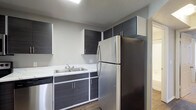 The kitchen is a standout feature of this single-floor space, showcasing a clean, modern aesthetic defined by dark textured cabinetry with sleek bar-pull hardware set against white cabinet frames, creating a striking two-tone contrast. Upper and lower cabinets provide ample storage, thoughtfully arranged to maximize functionality within the space. The countertops feature a light-toned surface that complements the dark cabinetry, while a double stainless steel sink adds a practical touch. Stainless steel appliances, including a full-size refrigerator and dishwasher, contribute to the kitchen's cohesive, contemporary look. The wood-look flooring anchors the space with warmth, balancing the cool tones of the cabinetry and appliances. Recessed and overhead lighting ensures the kitchen is well-lit, enhancing the overall brightness of the space. The neutral wall tones keep the design versatile and open, making the kitchen feel inviting and functional. Overall, the space reflects a modern design sensibility with practical, durable finishes suited for everyday living.
