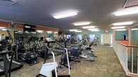 This single-floor fitness center offers a spacious and fully equipped environment designed to meet a wide range of exercise needs. The open layout accommodates an extensive collection of cardio machines, including treadmills, ellipticals, and stationary bikes, arranged to allow comfortable movement throughout the space. Additional strength and resistance training equipment lines the perimeter, providing users with a comprehensive workout experience. The walls are painted in warm terracotta and teal tones, giving the space an energetic and welcoming ambiance. Large mirrors are mounted along one wall, enhancing the sense of openness and allowing users to monitor their form during exercise. The textured ceiling features flush-mounted fluorescent lighting panels that illuminate the entire floor evenly. Patterned carpet covers the flooring throughout, offering a practical surface underfoot. Wall-mounted televisions are positioned for visibility across the room, adding an entertainment element to cardio sessions. Natural light filters in through windows near the far end of the space, complementing the overhead lighting. The overall design is functional and practical, prioritizing equipment accessibility and an open, motivating atmosphere suited for daily fitness use.