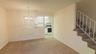 This 2-floor space presents a clean, functional layout with neutral tones throughout. The dining area is anchored by an elegant chandelier that adds a touch of warmth to the room, while soft, beige carpeting extends across the floor, creating a comfortable and cohesive atmosphere. A distinctive pass-through window connects the dining area to the kitchen, enhancing the sense of openness and allowing natural light to flow between the two spaces.

The kitchen is equipped with white cabinetry and matching white appliances, including a range and overhead ventilation hood, contributing to a bright and tidy aesthetic. The white cabinetry extends to the upper walls, maximizing storage capacity. Light-colored walls throughout maintain a consistent, airy feel across both rooms.

The textured ceiling adds subtle architectural character, while the straightforward layout ensures easy flow between areas. The pass-through window serves as a practical and visually interesting design element, bridging the kitchen and dining spaces. With its neutral palette and thoughtful layout, the space offers a versatile foundation suitable for a variety of interior styles.