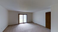 This three-floor property presents a straightforward and functional design throughout its multiple levels. The living area featured in the image is a spacious, open room finished with neutral wall-to-wall carpeting that contributes to a calm, understated atmosphere. The room is bathed in natural light courtesy of a full sliding glass door fitted with vertical blinds, which provides both privacy control and a direct view of the surrounding greenery outside. The sliding door also serves as a convenient point of access to the exterior.

The walls are finished in a clean, neutral white tone, keeping the space bright and versatile for various furniture arrangements. A floor-level heating vent is visible along the baseboard, indicating a standard forced-air climate system integrated into the flooring. The ceiling is smooth and white, maintaining a clean visual continuity throughout the room. The overall design of the living space is simple and unadorned, offering a blank canvas adaptable to a range of interior styles. With three floors in total, the property provides ample vertical living space to accommodate diverse functional needs across its levels.