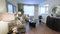 This single-floor space features a thoughtfully designed living area that combines comfort and functionality. The living room is anchored by warm hardwood-style flooring that runs throughout, complementing the soft yellow-toned walls that create an inviting, airy atmosphere. Large windows dressed with sheer white curtains and blinds allow generous amounts of natural light to fill the room, enhancing its open and bright character.

The living area is well-proportioned, comfortably accommodating a plush, cushioned sofa adorned with white accent pillows alongside a classic wingback chair, creating a cozy conversational arrangement. A dark oval coffee table serves as a central focal point, styled with decorative accents and greenery. Additional potted tropical plants are thoughtfully placed throughout, adding natural warmth and life to the space.

An open pass-through window connects the living area to an adjacent room, likely a kitchen, promoting a sense of openness and connectivity between spaces. Framed wall art and decorative wall accents contribute to the polished, well-curated aesthetic. Overall, the space presents a clean, comfortable, and inviting environment with a warm, neutral palette.
