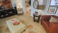 This two-floor residence offers a thoughtfully arranged living environment with a warm and welcoming ambiance. The main living area is anchored by light-toned hardwood flooring that flows seamlessly throughout, lending the space an airy, open feel. A plush ottoman-style coffee table sits atop a floral-patterned area rug, complementing the rich tones of a leather sofa and a classic wooden side table, which is topped with a modern lamp and subtle greenery. A patterned accent chair adds a charming touch of character to the room, while a bold oversized wall clock serves as a striking decorative focal point. Soft, ambient lighting from table lamps enhances the cozy atmosphere, while natural light filters in through slatted blinds. A hallway visible from the living area leads further into the home, hinting at additional rooms and living spaces across both floors. Decorative wall art featuring floral motifs adds a refined, homey quality to the interior. The overall design blends casual comfort with curated décor, creating a space that feels both lived-in and well-appointed. The two-floor layout provides ample room for versatile living arrangements.