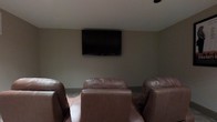 This dedicated media room is thoughtfully designed to deliver a focused and comfortable entertainment experience. The room centers around a large wall-mounted flat-screen television, positioned at an ideal viewing height to ensure clear sightlines from every seat. In front of the screen, a row of generously sized leather recliners provides ample seating, offering a plush and inviting place to relax during movie nights or extended viewing sessions. The neutral-toned walls and subdued ceiling contribute to a calm, distraction-free atmosphere that keeps attention on the screen. Overhead, a ceiling-mounted speaker or projector mount hints at enhanced audio-visual capabilities, suggesting the room is equipped to support a full home theater setup. The overall layout is intentional and efficient, maximizing the use of the space for its singular purpose. The muted color palette and controlled lighting environment further reinforce the cinematic ambiance, making this media room a standout feature ideal for entertainment enthusiasts seeking a private, theater-like retreat within the comfort of their own home.
