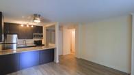 This single-floor space offers an open and functional layout designed for comfortable everyday living. The kitchen is a standout feature, outfitted with dark cabinetry, stainless steel appliances including a microwave and range, and a granite-style countertop that extends to form a convenient breakfast bar. The semi-open design of the kitchen allows it to connect naturally with the surrounding living area, creating a cohesive and social atmosphere.

Light wood-tone flooring runs throughout, lending warmth and continuity to the space. The walls are finished in a soft, neutral tone that enhances the brightness of the interior and complements the overall clean aesthetic. Overhead lighting provides practical illumination in the kitchen area.

A central hallway extends from the main living space, leading to additional rooms and offering a sense of depth and flow within the layout. The open corridor design ensures easy navigation throughout the unit, with doorways visible along the path suggesting well-proportioned secondary spaces.

Overall, the space strikes a balance between modern finishes and practical design, making it a well-appointed and inviting environment suited for a variety of living arrangements.