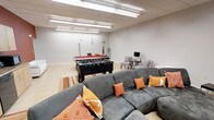 This single-floor recreational space is designed to accommodate both active gameplay and relaxed socializing. The room features a drop-panel ceiling with fluorescent strip lighting that provides bright, even illumination throughout the space. Light-toned tile flooring runs across the entire area, contributing to the open and clean aesthetic.

At the center of the room, a foosball table takes prominence, positioned in front of a full-sized pool table accompanied by a cue rack along the back wall. These gaming elements make the space well-suited for entertaining. A striped area rug anchors the seating zone, where a large, plush grey sectional sofa is adorned with orange and yellow accent pillows, offering generous seating for guests watching or waiting their turn.

Along one side of the room, a pair of zebra-print accent chairs flank a small side table with a lamp, creating a quieter conversation or reading nook. The walls are finished in a clean, neutral white, keeping the atmosphere bright and functional. Overall, the space strikes a balance between energetic recreation and comfortable lounging.
