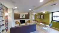 This single-floor space is a thoughtfully designed communal lounge that blends comfort with a lively, contemporary aesthetic. The room is anchored by a sleek fireplace set against a warm wood-paneled accent wall, above which hangs a striking moose portrait that adds a touch of rustic charm. Recessed ceiling lights and flush-mount fixtures provide bright, even illumination throughout the space, complementing the light hardwood flooring that extends across the entire area.

The lounge is divided into distinct functional zones. A relaxed seating area near the fireplace features a plush purple sofa paired with mustard-yellow accent chairs and natural wood stump side tables, creating a cozy gathering spot. On the opposite side, a dining and collaborative area offers dark wood tables paired with a mix of red, white, and yellow chairs, encouraging group interaction.

One wall is painted in a bold olive green and serves as a gallery wall, displaying a curated collection of framed prints that add color and personality to the space. The overall ambiance is energetic yet welcoming, with a carefully chosen color palette of earthy tones and vibrant accents that create a dynamic and engaging environment suitable for socializing, working, or unwinding.