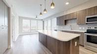 This well-designed one-bedroom, one-bathroom unit offers a thoughtfully organized layout that maximizes every square foot of its compact footprint. The open-concept kitchen and living area serves as the heart of the unit, featuring light wood-tone flooring that flows seamlessly throughout. The kitchen is outfitted with dark walnut-finished cabinetry, a geometric diamond-patterned tile backsplash, stainless steel appliances including a range, microwave, and French door refrigerator, and a generous curved island with a crisp white countertop. Three pendant lights with brass-toned fixtures hang above the island, adding a modern accent. The living area benefits from an expansive bank of windows fitted with horizontal blinds, filling the space with natural light and offering outdoor views. A ceiling fan serves the living zone. The bedroom is a comfortable, well-lit retreat with a large window, ceiling fan, and light-toned wood flooring. The bathroom features large-format tile flooring, a dark wood vanity with white countertop, a tiled shower-tub combination, and direct access to a dedicated closet with built-in shelving. Overall, the unit presents a clean, contemporary aesthetic throughout.