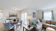This thoughtfully designed single-floor residence offers a cohesive and comfortable layout with a consistent neutral and teal color palette throughout. The open-concept living and dining area is anchored by light hardwood-style flooring, large sliding glass doors that invite natural light, and access to an outdoor space. The living room accommodates a generous sectional sofa and features multiple windows dressed with teal curtains, creating a bright and welcoming atmosphere. An adjacent dining area with a round wooden table completes the shared entertaining space. The kitchen is a standout feature, showcasing white shaker cabinetry, granite countertops, stainless steel appliances, and a large central island with bar seating illuminated by stylish pendant lighting — ideal for both cooking and casual dining. The bedroom is carpeted and well-lit by a broad window, offering a serene retreat. A secondary room with a closet serves flexibly as a home office, complete with plush carpeting and natural light. Two bathrooms are finished with tile flooring, white vanities, and clean, functional layouts — one featuring a shower-tub combination. Throughout, the space maintains a clean, modern aesthetic with warm accents and practical finishes.