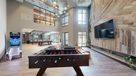 Located at 2801 Windy Ridge Pkwy SE in Smyrna, Georgia, this contemporary community amenity lounge is a well-appointed social hub designed for both relaxation and entertainment. The space is defined by its dramatic double-height ceilings in the main lounge area, which are adorned with striking sculptural wire chandeliers that serve as a bold focal point. Floor-to-ceiling steel-framed windows flood the interior with natural light and offer views of the surrounding outdoor areas.

The lounge zone features plush navy blue seating arranged around a foosball table, with a large-format TV mounted against a textured wood plank accent wall. An adjacent gaming area includes arcade machines and a shuffleboard table, while a colorful graffiti-style arcade cabinet and an eclectic mirror gallery wall add artistic personality to the space.

Transitioning into the social dining and bar area, the design shifts to a more structured aesthetic with exposed wood ceiling beams fitted with linear LED lighting, a large quartz-topped island, and rows of sleek bar-height tables with metal stools. A wet bar backed by brick and patterned tile provides a functional gathering point. Throughout, wide-plank wood-look flooring ties the entire space together, creating a warm, inviting, and thoroughly modern atmosphere ideal for community gatherings.