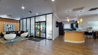 Located at 5800 Auvers Blvd in Orlando, Florida, this single-story community clubhouse offers a well-rounded set of amenities designed for both recreation and convenience. The spacious main lobby greets visitors with gleaming hardwood-style flooring, recessed lighting, and a sleek curved reception desk, creating a bright and professional first impression with views of the lush exterior through glass entry doors.

Adjacent to the lobby is a stylish lounge area furnished with a plush dark sectional sofa, modern egg-shaped chairs, and a colorful area rug — all positioned alongside floor-to-ceiling glass partition walls that provide a clear view into the dedicated fitness center. The fitness room, labeled "Family," is equipped with treadmills, exercise bikes, stability balls, and other workout equipment set against vibrant teal accent walls.

The game and entertainment room features warm orange accent walls, a full-size pool table, wall-mounted flat-screen TV, decorative guitar artwork, and a built-in counter workspace with seating — ideal for both leisure and productivity.

The full kitchen is outfitted with rich dark wood cabinetry, granite countertops, stainless steel appliances including a refrigerator, electric range, and microwave, and hardwood-style flooring throughout, offering a complete culinary space for community gatherings and events.