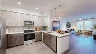 This thoughtfully designed one-bedroom, one-bathroom residence offers a functional and modern open-concept floor plan across a single level. The kitchen serves as a visual centerpiece, featuring two-tone cabinetry with light upper cabinets and warm-toned lower cabinets, white quartz countertops, stainless steel appliances, and a peninsula with pendant lighting that connects seamlessly to the dining and living areas. Wood-look plank flooring runs throughout the main living spaces, contributing to a cohesive and contemporary aesthetic.

The dining area accommodates a full table and benefits from proximity to both the kitchen and living room, making it well-suited for entertaining. The living room is bright and generously proportioned, with large windows and sliding glass door access to an outdoor area, allowing ample natural light throughout.

The bedroom features plush carpet, a ceiling fan, and a sliding glass door leading to a private balcony. The bathroom is finished with dark wood cabinetry, a white vanity surface, and a glass-enclosed walk-in shower. A defined entryway with closet access and utility connections provides a practical transition into the living space. Overall, the residence presents a clean, modern design with well-integrated finishes throughout.