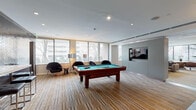 Located at 50 Main St in White Plains, NY, this single-floor amenity space is thoughtfully designed to balance recreation, work, and relaxation. The layout encompasses several distinct functional zones, each with its own character and purpose.

The central lounge and game area serves as a focal point, featuring a full-size billiards table set atop a striped wood-toned floor. Large windows along the perimeter flood the space with natural light and offer views of the surrounding landscape. A wall-mounted flat-screen television and modern lounge chairs complete this social hub. An adjacent seating area showcases a textured wood-paneled accent wall, a round white table surrounded by curved upholstered swivel chairs, and a decorative mirror — creating an intimate, stylish gathering spot.

A dedicated business center provides workstations with desktop computers, a printer, and a window view, supported by a separate conference room enclosed with glass-panel partitions. The kitchenette is equipped with warm wood cabinetry, a commercial-grade coffee brewer, and a stainless steel sink. A fitness center with exercise equipment is also present, along with accessible restrooms finished in neutral tile. Throughout, contemporary artwork and greenery contribute to a polished, welcoming atmosphere.