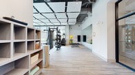 Located at 525 Lincoln St in Boston, MA, this contemporary fitness center offers a generously sized, single-floor layout designed to accommodate a full range of workout activities. The space makes a strong first impression with its sleek entry area, featuring bold black-framed glass walls and doors that provide visual connectivity throughout while defining distinct functional zones.

The main workout floor is outfitted with an impressive array of equipment, including a multi-station strength rack with a full dumbbell set, cable machines, adjustable benches, rowing machines, and a dedicated row of treadmills and stair climbers from premium brands. A large wall-to-wall mirror spans one side of the fitness floor, amplifying the sense of space and allowing users to monitor their form during training.

Overhead, a striking suspended ceiling grid of acoustic panels adds architectural interest while contributing to a comfortable acoustic environment. Exposed ductwork and pendant lighting complement the modern industrial aesthetic. Light-toned striped flooring runs throughout, lending warmth and continuity to the space.

Floor-to-ceiling windows along the perimeter flood the facility with natural light and offer views of the surrounding greenery, creating an energizing atmosphere. Storage shelving near the entrance provides convenient access to towels and amenities, rounding out this well-appointed, professional-grade fitness facility.