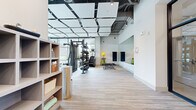 Located at 525 Lincoln St in Boston, MA, this contemporary fitness center offers a generously sized, single-floor layout designed to accommodate a full range of workout activities. The space makes a strong first impression with its sleek entry area, featuring bold black-framed glass walls and doors that provide visual connectivity throughout while defining distinct functional zones.

The main workout floor is outfitted with an impressive array of equipment, including a multi-station strength rack with a full dumbbell set, cable machines, adjustable benches, rowing machines, and a dedicated row of treadmills and stair climbers from premium brands. A large wall-to-wall mirror spans one side of the fitness floor, amplifying the sense of space and allowing users to monitor their form during training.

Overhead, a striking suspended ceiling grid of acoustic panels adds architectural interest while contributing to a comfortable acoustic environment. Exposed ductwork and pendant lighting complement the modern industrial aesthetic. Light-toned striped flooring runs throughout, lending warmth and continuity to the space.

Floor-to-ceiling windows along the perimeter flood the facility with natural light and offer views of the surrounding greenery, creating an energizing atmosphere. Storage shelving near the entrance provides convenient access to towels and amenities, rounding out this well-appointed, professional-grade fitness facility.
