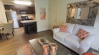 This single-floor residence offers a well-designed open-concept layout that seamlessly connects the living area to the kitchen, creating a cohesive and functional living environment. The living room is warm and inviting, featuring hardwood-style flooring that extends throughout the space, complemented by neutral green-toned walls that lend a calm, airy feel. The room accommodates comfortable seating and is styled with tasteful accent pieces, including an ornate wall mirror and colorful throw pillows that add pops of orange to the neutral palette. A rectangular coffee table anchors the seating area, providing both style and practicality. Adjacent to the living space, the kitchen is outfitted with rich dark cabinetry, light granite-style countertops, and black appliances, including a range and built-in microwave. A kitchen island with seating offers additional prep space and a casual dining option. The entry door is conveniently located off the living area, providing a natural transition between the interior and exterior. Overall, the space strikes a balance between modern finishes and cozy, residential charm, making it both functional and aesthetically appealing.