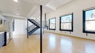 Located in the heart of Downtown Los Angeles on South Broadway, this two-bedroom, two-bathroom two-story loft blends industrial character with modern design. The main floor is expansive and open, anchored by light bamboo-style hardwood flooring that runs throughout the space. A dramatic steel staircase with metal-grate treads and horizontal rail balusters connects the two levels, serving as a striking architectural focal point. Exposed ductwork overhead and soaring ceilings reinforce the industrial loft aesthetic, while recessed lighting provides a clean, contemporary touch. The kitchen features blue lower cabinetry, white upper cabinets with a subway tile backsplash, and stainless steel appliances, including a refrigerator and range. The upper level offers an open loft area bathed in natural light from large steel-framed windows with city views, providing a versatile secondary living or workspace. The two bedrooms are bright and minimalist, with large windows fitted with zebra-style roller shades. The bathrooms offer distinct designs: one features frosted glass enclosures and a sleek, modern finish, while the other includes a tub-shower combination with white subway tile, mosaic accent tile, and dark slate-look floor tile. The space balances raw urban character with refined finishes throughout.
