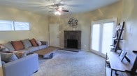 This single-story space centers around a comfortable living room designed for both relaxation and functionality. The room is anchored by a brick fireplace with a raised hearth, serving as a natural focal point and adding warmth and character to the space. Plush wall-to-wall carpeting covers the floor, contributing to a cozy, inviting atmosphere, while neutral-toned walls provide a versatile backdrop throughout.

A ceiling fan with an integrated light fixture is centrally positioned, offering both air circulation and ambient lighting. French doors along one wall allow natural light to filter into the room, brightening the interior and providing easy access to the outdoors. A standard interior door provides additional access points within the home.

The living area comfortably accommodates a seating arrangement, including a sofa and accent chair, along with a coffee table, demonstrating the room's generous proportions. Decorative wall art adds a personal touch above the fireplace. Overall, the space presents a functional and welcoming single-floor layout with classic residential features throughout.