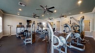 This fitness center is organized across a single floor and divided into distinct functional zones designed to support a full range of workout activities. The main exercise area features durable dark rubber flooring and hosts an extensive selection of equipment, including treadmills, elliptical trainers, stationary and recumbent bikes, and a variety of cable-based strength training machines. Multiple ceiling fans are mounted throughout, complementing the recessed lighting to maintain a comfortable environment during workouts. A wall-mounted television provides entertainment options for users on cardio equipment. Natural light enters through several windows along the perimeter walls, adding brightness to the space. The walls are finished in neutral tones with white crown molding and chair rail details, lending a clean, polished appearance. A separate studio room adjoins the main floor and is finished with patterned carpet tiles. This room is equipped with stability balls, resistance accessories, a dumbbell rack, and a floor-to-ceiling mirror along one wall, making it well-suited for stretching, group fitness, or individual training sessions. A large interior window connects the studio to the main area, maintaining visual openness between the two spaces. An exterior door in the studio provides direct outdoor access.