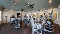 This fitness center is organized across a single floor and divided into distinct functional zones designed to support a full range of workout activities. The main exercise area features durable dark rubber flooring and hosts an extensive selection of equipment, including treadmills, elliptical trainers, stationary and recumbent bikes, and a variety of cable-based strength training machines. Multiple ceiling fans are mounted throughout, complementing the recessed lighting to maintain a comfortable environment during workouts. A wall-mounted television provides entertainment options for users on cardio equipment. Natural light enters through several windows along the perimeter walls, adding brightness to the space. The walls are finished in neutral tones with white crown molding and chair rail details, lending a clean, polished appearance. A separate studio room adjoins the main floor and is finished with patterned carpet tiles. This room is equipped with stability balls, resistance accessories, a dumbbell rack, and a floor-to-ceiling mirror along one wall, making it well-suited for stretching, group fitness, or individual training sessions. A large interior window connects the studio to the main area, maintaining visual openness between the two spaces. An exterior door in the studio provides direct outdoor access.