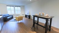 This well-appointed single-floor space offers an inviting open-concept layout that combines living and dining areas into one cohesive environment. Rich hardwood flooring flows throughout, adding warmth and a sense of continuity to the space. Large windows along the exterior wall flood the interior with generous natural light, creating a bright and airy atmosphere while offering views of the surrounding greenery outside.

The living area is thoughtfully arranged with comfortable seating, including a pair of mustard-yellow accent chairs positioned on a textured area rug, anchoring the lounge zone. A low media console serves as a functional focal point in this area, while a piece of artwork leans against the wall, adding a casual, curated touch to the space.

Adjacent to the living area, a tall bar-height dining table with industrial-style adjustable stools defines the dining zone, offering a stylish and practical space for casual meals or gathering. The neutral, light-toned walls provide a clean backdrop that complements the dark furniture accents throughout.

The overall ambiance is modern yet relaxed, with an emphasis on open, uncluttered living. The thoughtful combination of natural light, warm flooring, and contemporary furnishings makes this space feel both functional and welcoming.