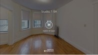 This single-floor studio offers a thoughtfully arranged layout that maximizes its compact footprint. The main living area is highlighted by a angled bay window configuration fitted with roller shades, allowing for controlled natural light while providing views of the surrounding urban streetscape. Warm-toned hardwood floors run throughout the main living space, lending a classic character to the interior. A cast-iron radiator positioned near the bay window provides traditional heating, a nod to the building's older architectural roots. The textured ceiling adds additional period detail to the space.

The separate kitchen features tile flooring, white cabinetry, a gas range, a full-size refrigerator, and a sink with countertop workspace. A doorway connects the kitchen directly to the main entry, creating a practical flow through the unit. An additional smaller room off the main living area offers flexible use as a sleeping nook or storage space.

The full bathroom includes a tub-shower combination with a curtain rod, a white vanity with under-sink storage, and blue ceramic tile accents along the walls, complemented by neutral tile flooring. Overall, the space reflects a functional, classic urban character.