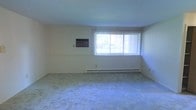This single-floor residential space offers a clean, functional layout designed for comfortable everyday living. The main living area is a standout feature, characterized by its generous proportions and neutral, light-toned walls that create a calm and welcoming ambiance. A large window fitted with vertical blinds allows an abundance of natural light to fill the room, enhancing the open and airy feel of the interior. The floor is finished with wall-to-wall carpeting in a soft, neutral tone, adding warmth and comfort underfoot. A baseboard heating system runs along the exterior wall beneath the window, ensuring efficient climate control throughout the colder months. A wall-mounted panel, likely a thermostat or intercom unit, is positioned conveniently within the living space. The overall design is straightforward and practical, with smooth ceilings and uncluttered wall surfaces that provide a versatile backdrop for a variety of furnishing styles. The space presents a solid foundation for personalization, offering a quiet, well-lit environment suitable for a range of living arrangements.