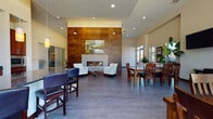 This single-floor community facility is thoughtfully designed to offer a variety of gathering and amenity spaces within a contemporary architectural framework. The main clubroom serves as the centerpiece, anchored by a dramatic floor-to-ceiling wood-paneled wall housing a sleek linear gas fireplace. A pair of accent chairs flanks the hearth, creating an intimate seating area, while nearby dining tables accommodate larger groups. Large floor-to-ceiling windows flood a dedicated dining zone with natural light and views of the surrounding exterior, complemented by a wall-mounted television for entertainment.

A fully equipped kitchen sits adjacent to the social areas, featuring rich dark wood cabinetry, granite countertops, stainless steel appliances including a refrigerator, dishwasher, and built-in microwave, and tile flooring bathed in natural light from a side window.

The fitness center is well-appointed with cardio equipment including treadmills, ellipticals, and stationary bikes, rubber flooring, full-wall mirrors, and vibrant accent walls. Natural light enters through multiple windows, enhancing the energetic atmosphere.

Entry foyers on both sides of the facility feature soaring ceilings, hardwood-style flooring, and glass entry doors that draw in daylight, creating a welcoming first impression throughout.