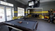 This single-floor game room is designed for entertainment and social interaction, featuring a striking black and yellow color scheme that sets an energetic tone throughout the space. Bold yellow horizontal stripes accent the dark gray walls, complemented by a black and yellow checkered floor detail near the dining area, creating a cohesive and dynamic aesthetic.

At the center of the room sits a full-size pool table with a slate-blue felt surface, ready for play and serving as the focal point of the space. A foosball table nearby adds another layer of recreational options. Overhead, a ceiling-mounted multi-screen TV setup provides excellent sightlines from virtually any point in the room, making it ideal for watching sports or gaming.

Natural light filters into the space through large windows, offering views of mature trees and outdoor greenery, balancing the bold interior atmosphere with a refreshing connection to the outdoors. Crown molding along the ceiling adds a refined architectural detail that elevates the room's overall finish.

A dedicated dining and card-game area is furnished with a dark table and cushioned chairs, providing a comfortable setting for group activities. The layout is open and well-organized, making the space functional for a variety of social gatherings and recreational activities.