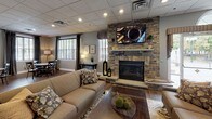 This single-floor community facility is thoughtfully designed to offer residents a comfortable and functional gathering space. The grand entrance lobby sets an inviting tone with soaring ceilings, decorative beam accents, textured wallcovering, and rich wood-look tile flooring that transitions seamlessly throughout the space. A chandelier and wall-mounted sconces provide warm, layered lighting, while a console table with a flat-screen TV anchors the entryway. Adjacent to the lobby is a relaxed seating nook featuring upholstered wingback chairs, wall art, and lush greenery — ideal for quiet conversation. The lounge area centers around a dramatic stacked-stone fireplace with a wall-mounted television above, complemented by plush seating, hardwood-style flooring, and large windows that bring in natural light. The fully equipped kitchen boasts white shaker cabinetry, granite countertops, a subway tile backsplash with decorative accent band, stainless steel appliances, and an eye-catching stacked-stone kitchen island with bar seating. The fitness center is outfitted with a variety of cardio and strength equipment set against vibrant teal accent walls, providing an energizing workout environment. Together, these spaces create a well-rounded, amenity-rich facility.