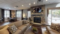This single-floor community facility is thoughtfully designed to offer residents a comfortable and functional gathering space. The grand entrance lobby sets an inviting tone with soaring ceilings, decorative beam accents, textured wallcovering, and rich wood-look tile flooring that transitions seamlessly throughout the space. A chandelier and wall-mounted sconces provide warm, layered lighting, while a console table with a flat-screen TV anchors the entryway. Adjacent to the lobby is a relaxed seating nook featuring upholstered wingback chairs, wall art, and lush greenery — ideal for quiet conversation. The lounge area centers around a dramatic stacked-stone fireplace with a wall-mounted television above, complemented by plush seating, hardwood-style flooring, and large windows that bring in natural light. The fully equipped kitchen boasts white shaker cabinetry, granite countertops, a subway tile backsplash with decorative accent band, stainless steel appliances, and an eye-catching stacked-stone kitchen island with bar seating. The fitness center is outfitted with a variety of cardio and strength equipment set against vibrant teal accent walls, providing an energizing workout environment. Together, these spaces create a well-rounded, amenity-rich facility.