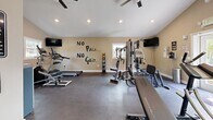 Located at 701 E Bay Dr in Largo, Florida, this dedicated fitness center offers a well-organized and fully equipped exercise space designed to support a comprehensive workout routine. The room features vaulted ceilings that lend an open, airy feel, complemented by ceiling fans and a wall-mounted mini-split unit to maintain a comfortable environment during workouts. Natural light filters in through multiple windows, brightening the warm, neutral-toned walls adorned with motivational typography decals, including the iconic "No Pain... No Gain" and "Eat. Sleep. Lift. Repeat." phrases. The dark epoxy flooring provides a durable, easy-to-maintain surface throughout. Equipment includes a treadmill, elliptical, stationary bike, and a multi-station cable machine for cardio and strength training. A full dumbbell rack with an adjustable weight bench serves free weight enthusiasts, while a medicine ball rack adds versatility. Wall-mounted flat-screen TVs on opposite ends of the room offer entertainment during sessions. A large wall mirror enhances the sense of space and allows for proper form monitoring. Additional amenities include a water dispenser, a fire extinguisher, and posted facility rules, reflecting attention to safety and user convenience. A single entry door provides straightforward access to this thoughtfully appointed fitness facility.
