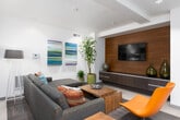 This single-floor space is thoughtfully designed with a contemporary aesthetic, seamlessly integrating a living area and kitchen within an open, functional layout. The living area serves as the centerpiece, anchored by a spacious sectional sofa in a neutral grey fabric with multicolored accent pillows. A warm wood coffee table sits at its center, complementing the rich wood-paneled feature wall, which houses a flat-screen television and a sleek, low-profile media console. Vibrant abstract canvas artwork in shades of blue, green, and purple adds a bold visual element to the white walls, while tall potted tropical plants introduce natural texture and life to the space.

The kitchen is efficiently appointed with dark espresso cabinetry extending floor to ceiling, light-toned countertops, and a blue tile backsplash that adds a subtle pop of color. Stainless steel appliances, including a refrigerator and dishwasher, provide a polished, modern finish. Large-format light tile flooring flows throughout, lending a clean, cohesive feel.

An adjacent secondary seating area features a striking orange accent chair and a bistro-height table with white bar stools set against a bold teal accent wall, adding energy and visual contrast. Recessed lighting throughout ensures a well-lit, inviting ambiance.