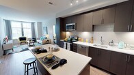 This single-floor residence showcases a thoughtfully designed open-concept layout where the kitchen and living area interact seamlessly. The kitchen serves as a central hub, anchored by a generous island with a clean white countertop that doubles as a breakfast bar with seating. Rich, dark-toned cabinetry extends from lower to upper sections, providing substantial storage while lending a sophisticated, contemporary aesthetic. Stainless steel appliances — including a built-in microwave, electric range, and dishwasher — complement the modern design, while a sleek faucet and undermount sink add functional elegance. A white subway tile backsplash provides a clean contrast against the darker cabinetry. Recessed lighting illuminates the kitchen with precision, highlighting the well-appointed surfaces throughout. The space opens naturally into a bright living area, where large windows allow natural light to fill the room, creating an inviting and airy atmosphere. Light wood-style flooring runs throughout, unifying the open floor plan cohesively. The overall ambiance is modern and polished, balancing functionality with refined residential design.
