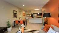 This single-floor residence showcases a thoughtfully designed open-concept layout, where the kitchen, dining, and living areas flow together to create a cohesive and functional living environment. The kitchen serves as a central focal point, featuring a large white waterfall-edge island with an integrated sink, dark espresso cabinetry, stainless steel appliances, and stylish pendant lighting that hangs above the countertop. Open floating shelves provide both storage and display space, adding a decorative touch to the kitchen zone. Light wood-style flooring runs throughout the main living area, contributing to a clean and modern aesthetic. The dining area comfortably accommodates a full-sized table and chairs, positioned adjacent to the kitchen for convenient everyday use. A bold accent wall adds a warm pop of color to the living space, complementing the neutral tones found throughout the rest of the unit. Recessed lighting and pendant fixtures ensure well-distributed illumination across the open floor plan. The overall ambiance is contemporary and welcoming, with a cohesive design palette of neutral walls, dark cabinetry, and warm accents creating a balanced and inviting atmosphere.